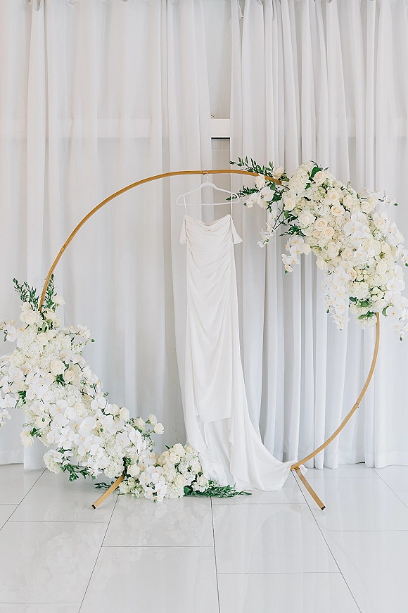 Wedding dress display on a hanger suspended from a gold circular arch with white roses and orchids against white draped curtains indoors