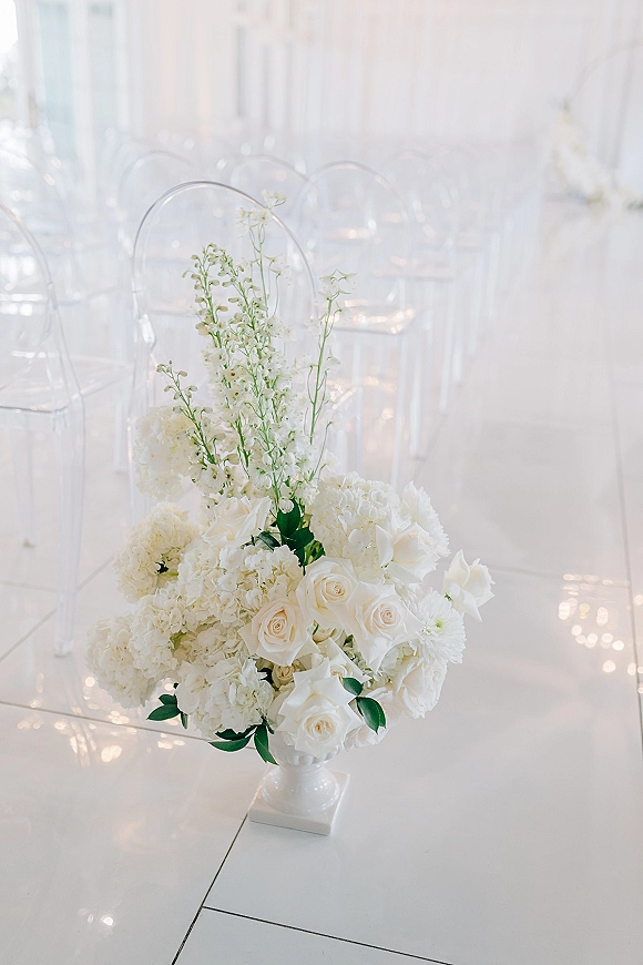Ceremony aisle flowers in a white pedestal vase with white roses, hydrangeas, and greenery beside a white runner and clear chairs