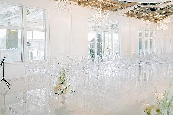 Indoor ceremony setup with clear acrylic chairs lining a wide aisle, white rose urns and chandeliers beneath flowing white draping in a bright room