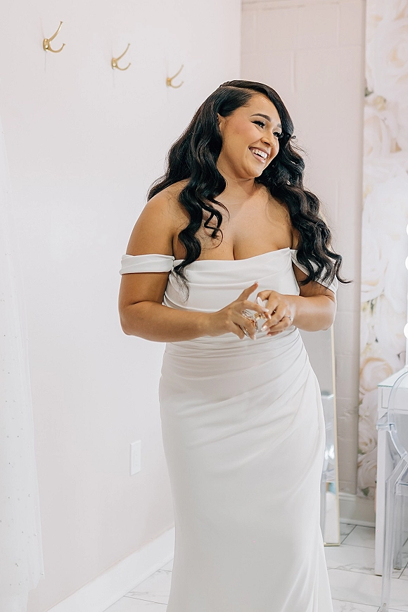 Bridal portrait of a getting ready bride laughing in an off the shoulder satin wedding dress, showing her ring in a bright suite with mirror