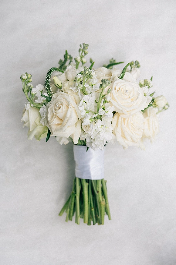 Bridal bouquet of white roses with greenery, filler flowers, and satin ribbon wrap with pearl pins against a light gray backdrop