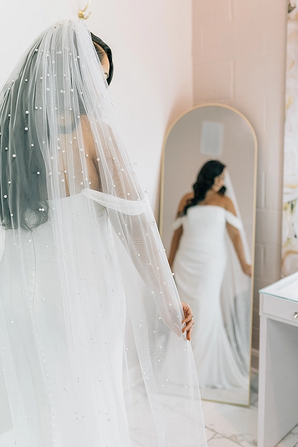 Bridal portrait of a bride in mirror wearing a pearl wedding veil and off the shoulder dress in a light pink bridal suite with vanity