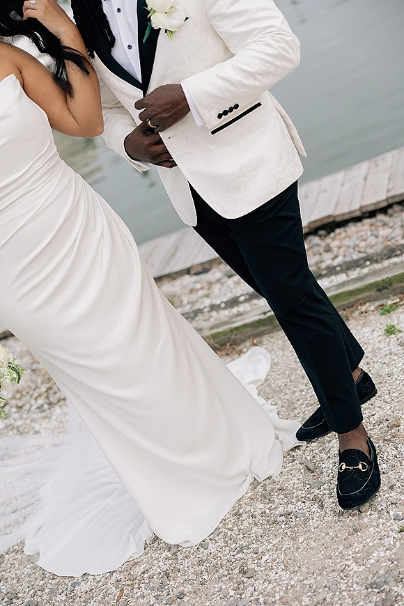 Couple portrait of bride holding groom’s lapel, engagement ring visible, by a dock with water behind, her strapless gown train flowing