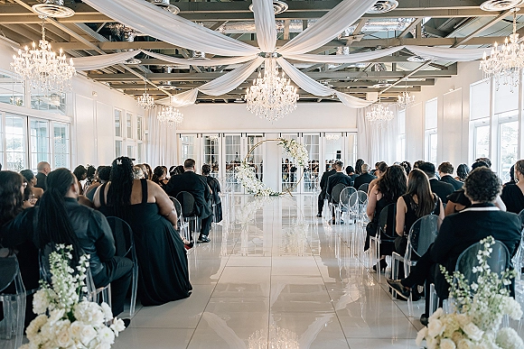 Indoor wedding ceremony with crystal chandeliers and a circular arch, white floral aisle arrangements, and clear acrylic chairs for seated guests