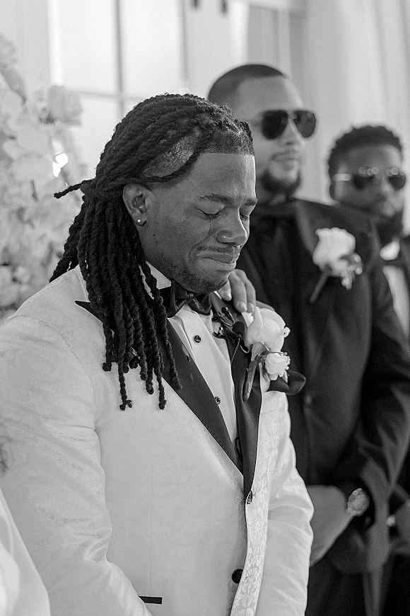 Groom portrait capturing an emotional groom moment in a white tuxedo with black bow tie and boutonniere, guests and florals behind in window light
