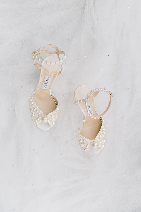Bridal shoes with pearl wedding heels, ankle straps, and open toes resting on tulle with scattered pearls against a white backdrop