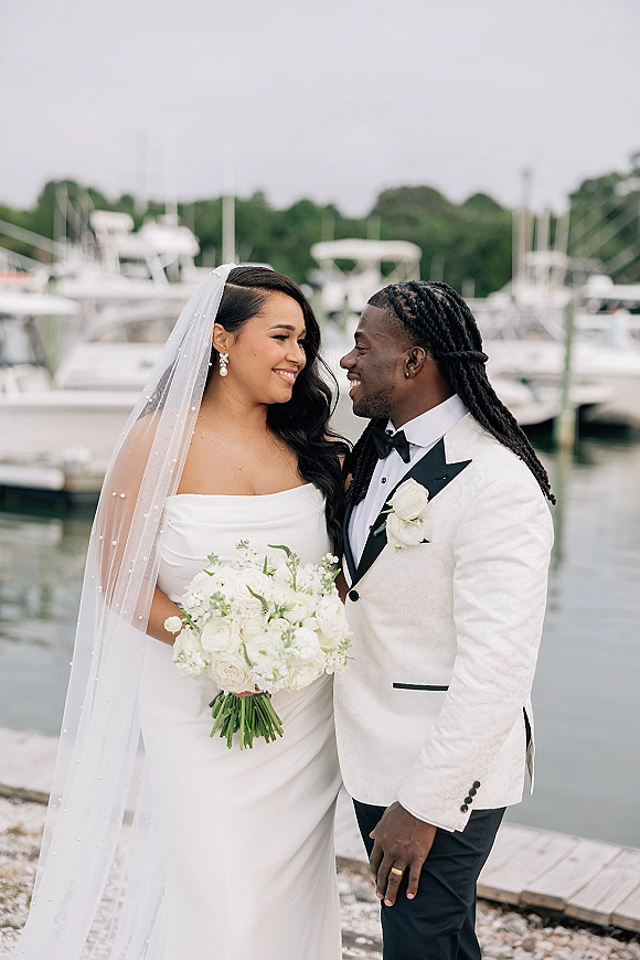 Couple portrait of bride in strapless dress and veil holding a white bouquet beside groom in bow tie at an overcast marina dock