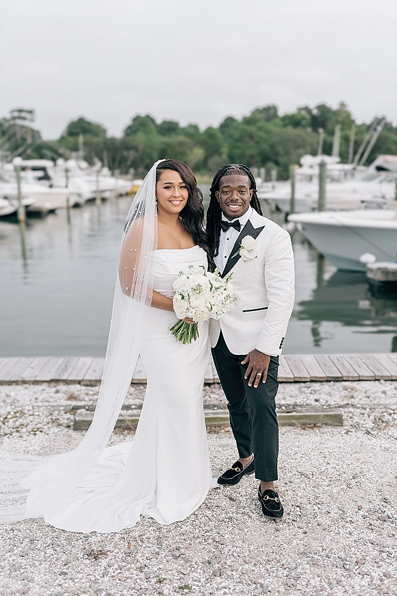 Couple portrait of bride and groom smiling, bride holding white bouquet, by a marina dock with boats and water under overcast sky