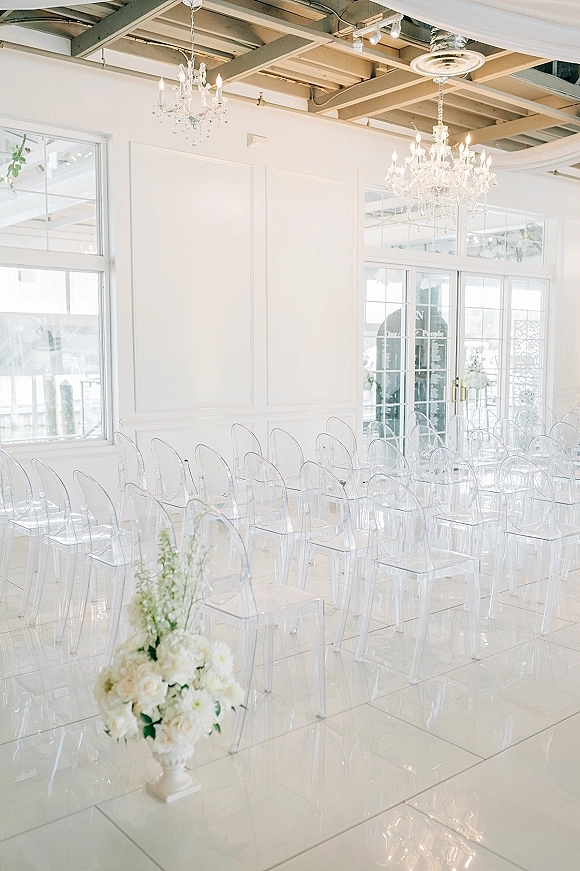 Indoor ceremony setup with clear acrylic ceremony chairs facing a white floral urn beneath crystal chandeliers in a bright white hall