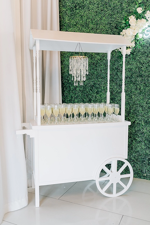 Champagne cart wedding champagne cart setup with flutes and bottles on a white bar cart beneath a chandelier, against a boxwood hedge wall