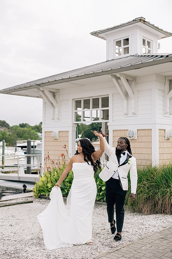 Couple portrait of bride and groom walking hand in hand on a waterfront dock, her long veil trailing by boats and a white boathouse