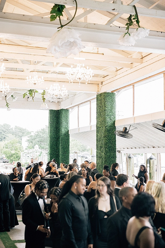 Wedding reception with guests mingling at cocktail hour under white chandeliers, hanging flowers, and greenery garlands in a bright pavilion