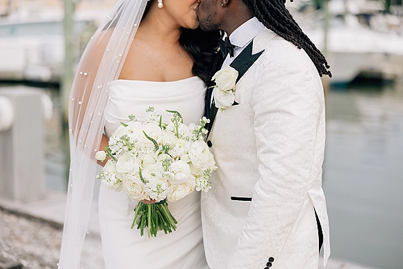 Wedding kiss as bride holds a white rose bouquet, veil and pearls framing the couple in tuxedo by a marina dock railing and boats