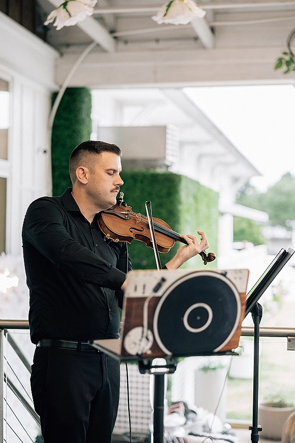 Wedding musician playing violin with bow beside a music stand on a white porch, green hedges behind, dressed in a black shirt