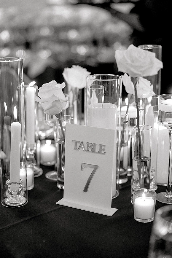Wedding table centerpiece with white roses and pillar candles beside an acrylic table number 7, set on a tabletop with blurred reception tables