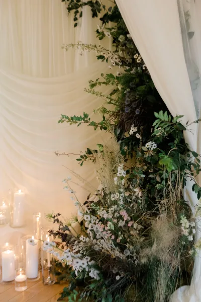 Wedding ceremony backdrop with greenery ceremony backdrop, white draping, wildflowers, pampas grass, and pillar candles on a wooden floor