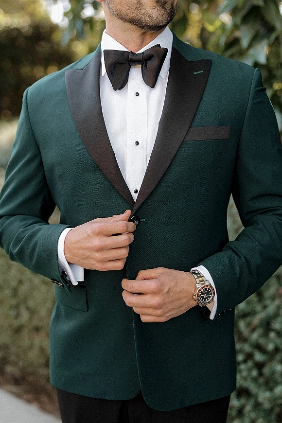 Groom tuxedo details with emerald green jacket, black shawl lapel, and bow tie, showing cufflinks and wristwatch against blurred greenery