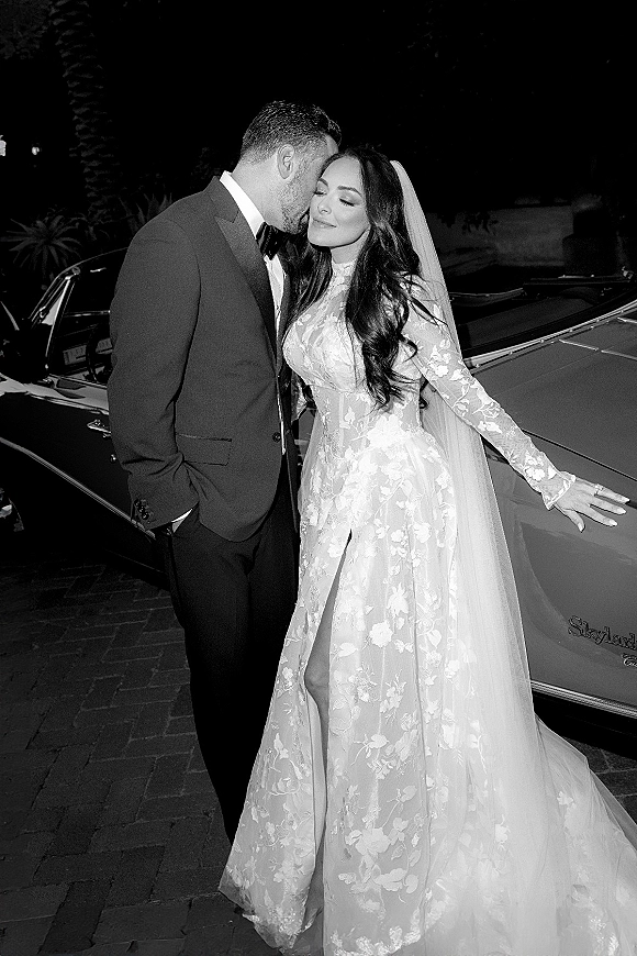 Couple portrait, black and white wedding photo of groom kissing bride’s forehead beside a vintage convertible on a palm-lined driveway at night