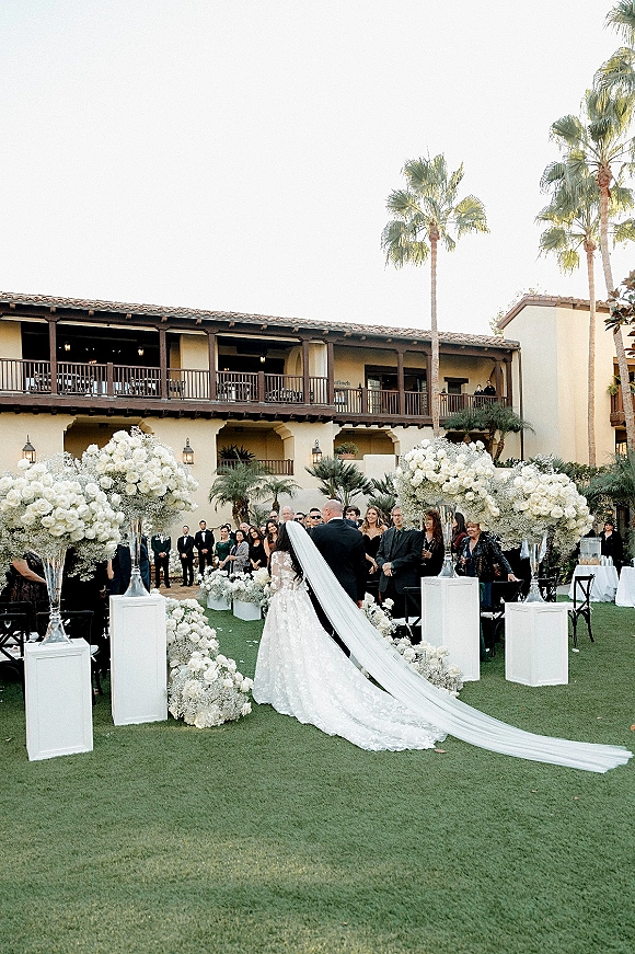 Ceremony processional as bride walking down aisle in lace gown with long veil, passing white flowers on pedestals in Spanish courtyard lawn