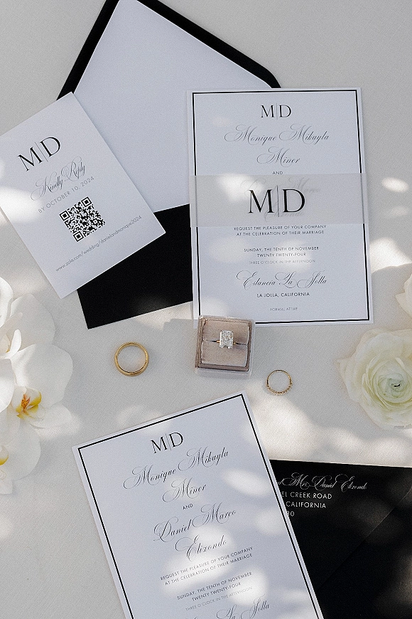 Invitation suite with black and white wedding invitations, RSVP and QR code cards, rings, and white roses on a white surface with soft shadows