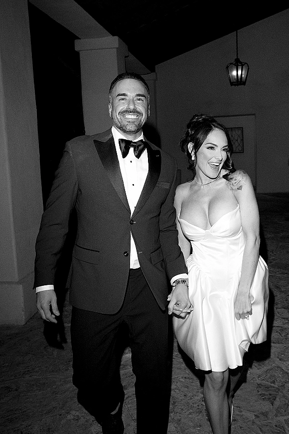 Newlywed couple in a wedding exit photo walking hand in hand at night through a stucco corridor with columns and a lantern backdrop