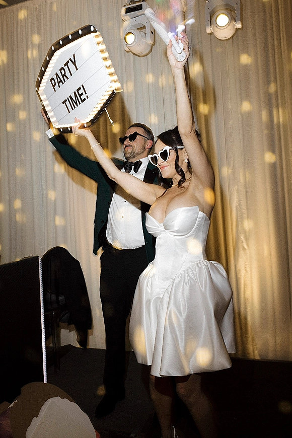 Wedding reception party with newlyweds dancing in heart sunglasses as the bride pops confetti on a disco-lit dance floor by draped curtains