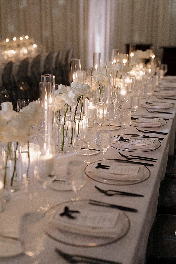 Reception tablescape with a long table wedding setup, hurricane candles, taper candles, white florals, and black ribbon menus in a dim draped room