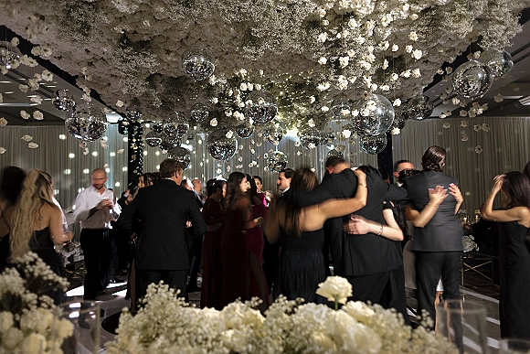 Wedding dance floor packed with guests dancing beneath hanging floral ceiling of white roses and baby's breath, disco balls, and string lights in a ballroom