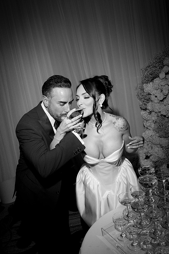 Wedding toast moment as newlyweds drinking champagne from coupe glasses beside a champagne tower table, framed by draped curtains indoors