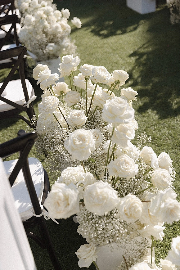 Ceremony aisle flowers with white roses and baby's breath, tied with white ribbon on black chairs along a grass lawn outdoor aisle