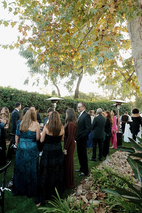 Wedding cocktail hour with outdoor cocktail hour guests mingling around cocktail tables, holding drinks on a fall garden lawn by patio heaters