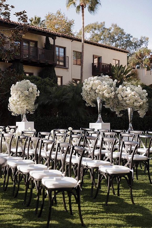 Ceremony setup with black folding chairs and white rose arrangements lining the aisle on a lawn in front of a villa with palm trees.