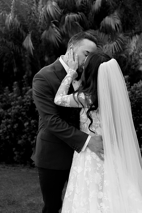 Wedding kiss portrait of bride and groom kissing as she cups his face, lace dress and veil beside his tuxedo with lush tropical greenery behind