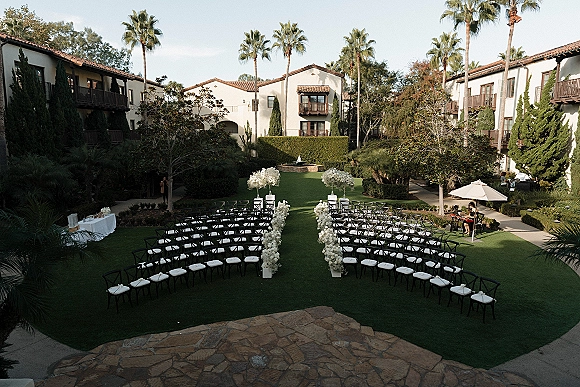 Ceremony setup with white floral aisle markers and black chairs on a courtyard lawn, lined with palms and a fountain backdrop