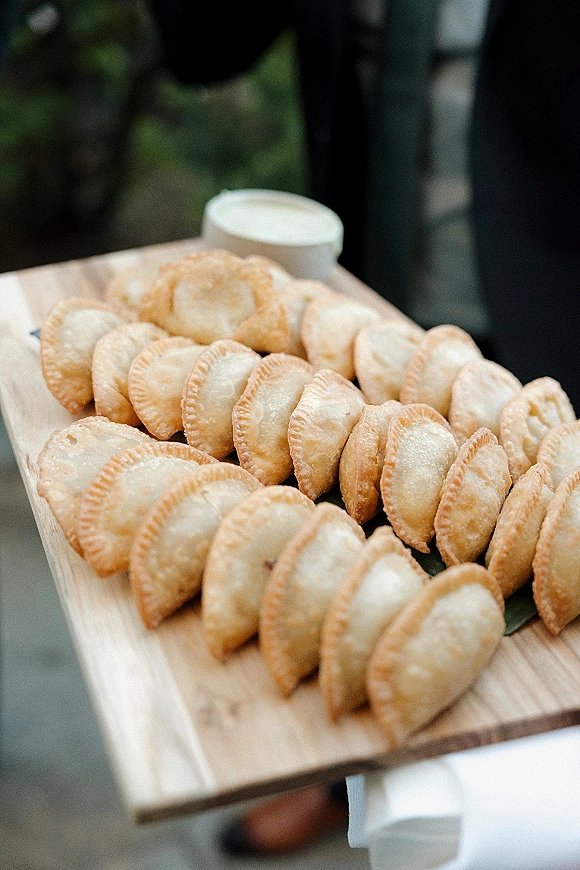 Wedding appetizers on a wood serving board with fried pastry bites and a dipping sauce cup, held by a guest on an outdoor walkway with greenery
