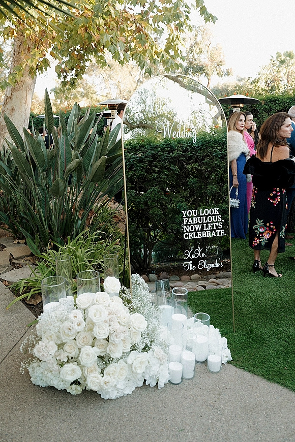 Wedding welcome sign on an arched mirror with calligraphy lettering, white roses and hydrangeas, flanked by pillar candles on a garden patio walkway