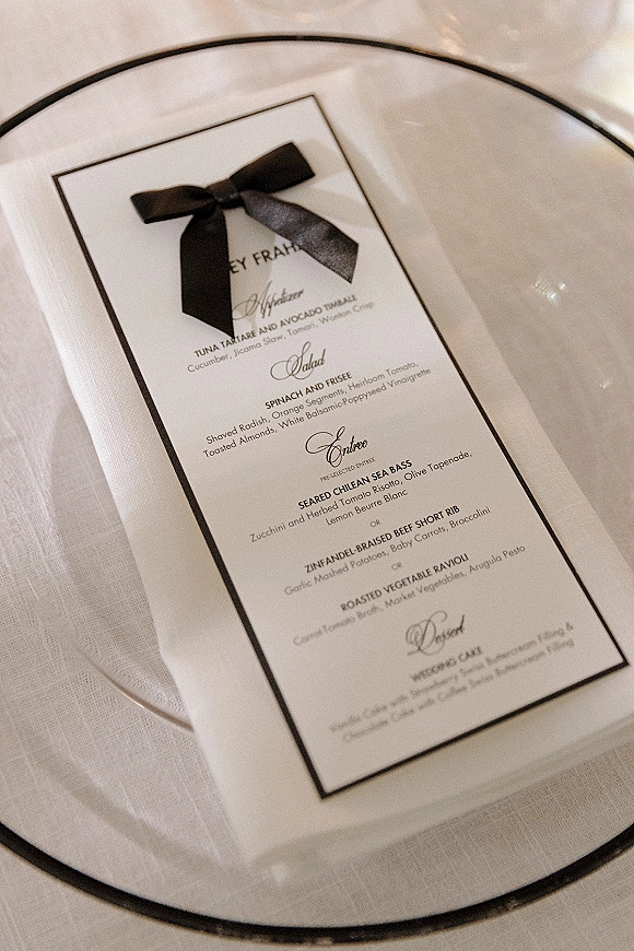 Wedding menu card with black satin ribbon bow on a black-rim dinner plate, resting on a white linen napkin over a white tablecloth