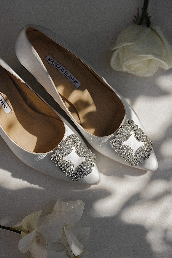 Bridal shoes in white satin with crystal buckle detail resting on white fabric, paired with a rose and orchid in soft natural light