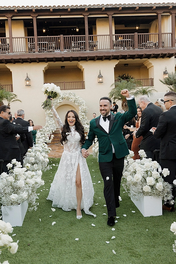 Wedding recessional as bride and groom hold hands, she lifts bouquet overhead while guests toss petals in a palm-lined courtyard aisle