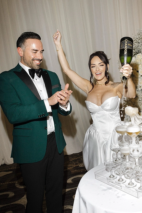 Champagne tower moment as bride in strapless gown and groom in tux pour champagne into coupe glasses, cascade over white tablecloth.