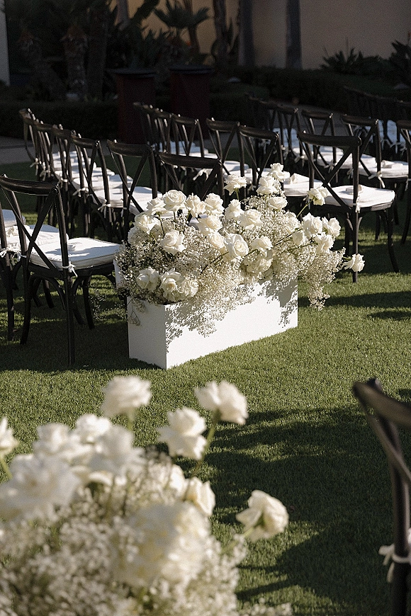 Ceremony aisle flowers in white rose aisle decor along a grass lawn, with baby's breath boxes beside black crossback chairs in a garden setting