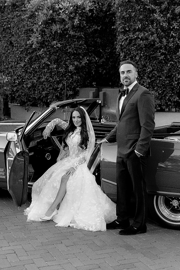 Couple portrait of bride in a wedding dress and veil in a vintage convertible car, groom in tuxedo with bow tie on driveway by hedges