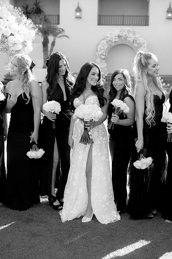 Bridesmaids portrait with bride and bridesmaids in black bridesmaid dresses holding white rose bouquets on a lawn by a stucco facade and palms
