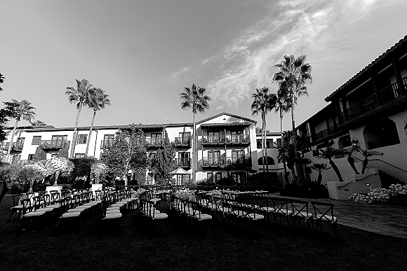 Ceremony setup for an outdoor wedding ceremony with black crossback chairs and a flower-lined aisle on a palm-lined Spanish-style courtyard lawn