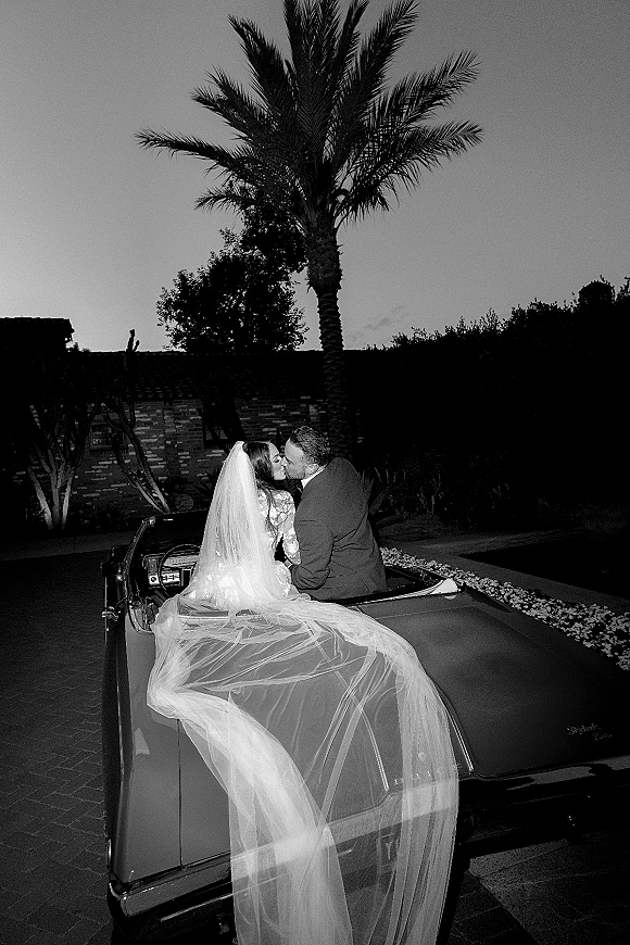 Wedding kiss portrait of newlyweds in a convertible, bride’s veil draped over the vintage car under palm trees at night