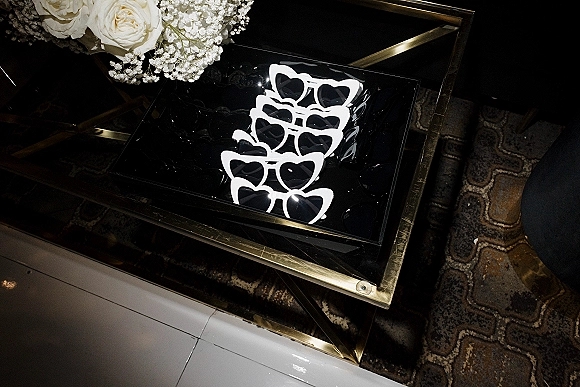 Wedding sunglasses in a gold-framed glass display box with white heart frames and roses on patterned carpet and tiled floor
