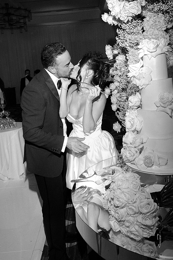 Wedding cake cutting as bride in strapless dress and groom in tux slice a tiered cake with rose topper beside champagne coupe tower indoors