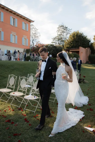 Newlywed portrait of bride and groom walking arm in arm with champagne glasses, veil blowing near villa lawn seating and guests
