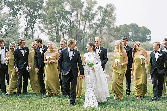 Wedding party portrait of bride and groom with bridal party walking on a grass lawn, bridesmaids in olive green dresses holding white bouquets