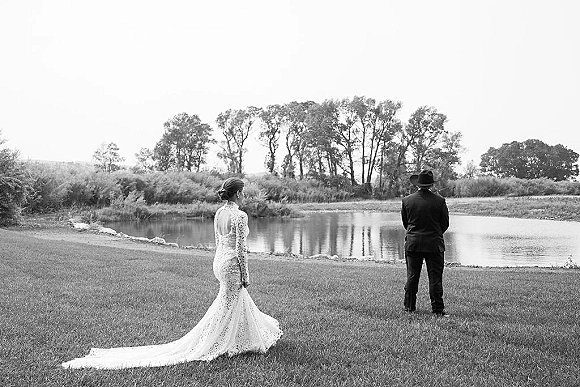Wedding first look as bride in a lace long train dress walks up behind groom in a cowboy hat by a lakeside pond with trees